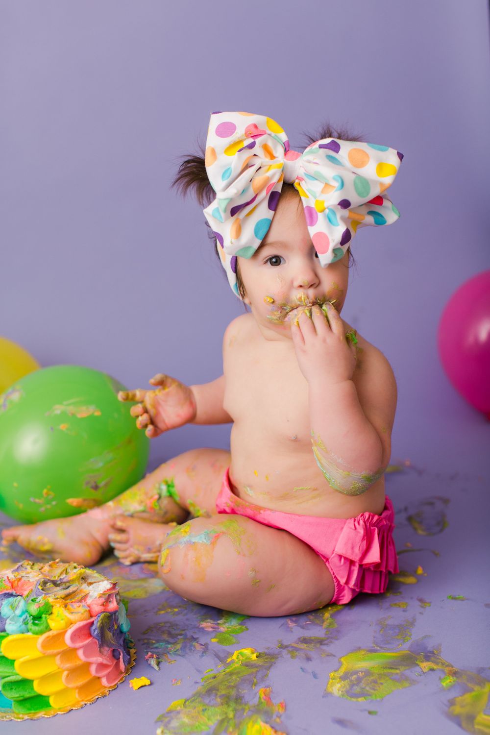 Baby enjoying messy cake smash photoshoot with colorful balloons and rainbow cake on purple backdrop.