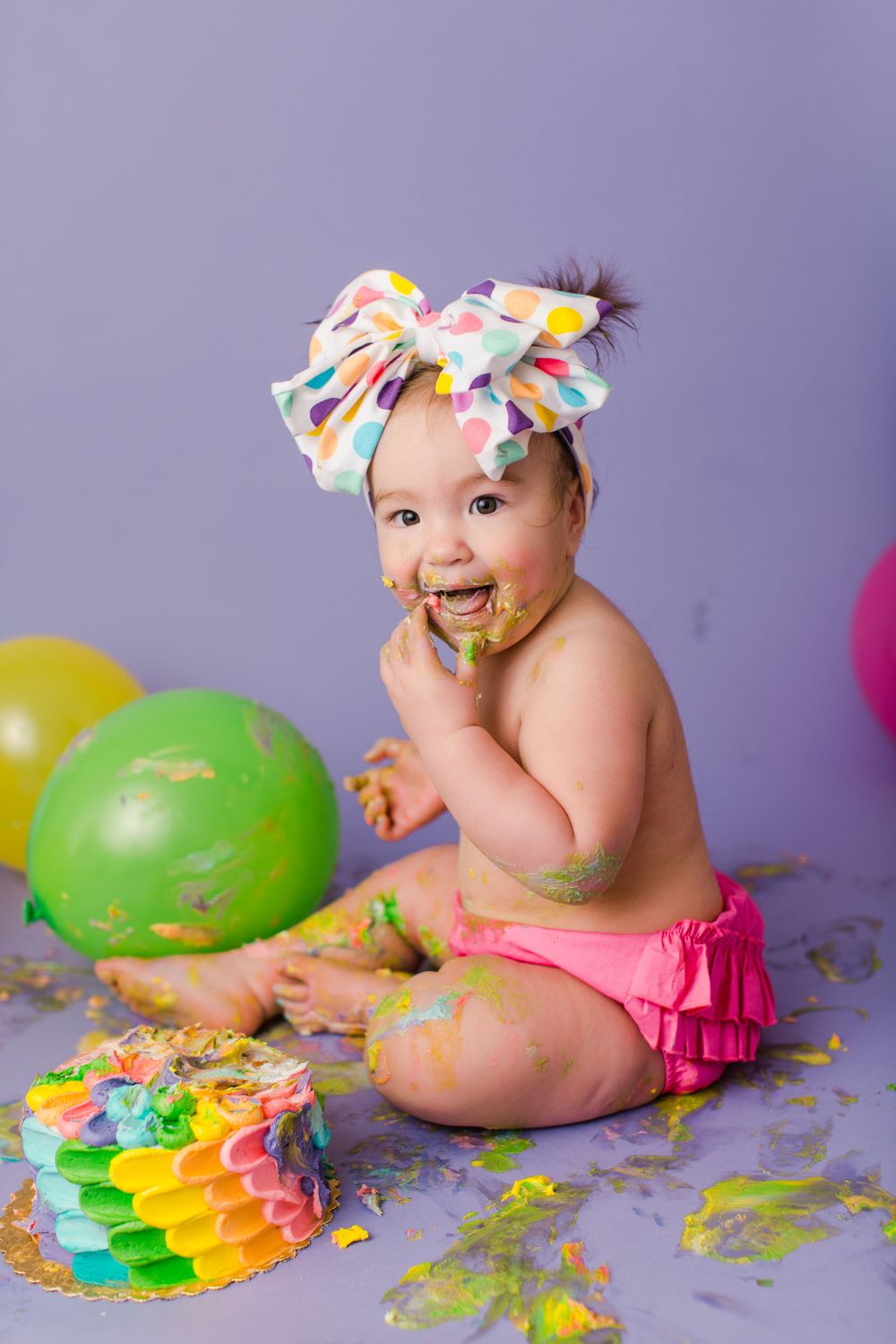 Baby wearing large polka dot bow plays with green balloon during festive cake smash photography session.