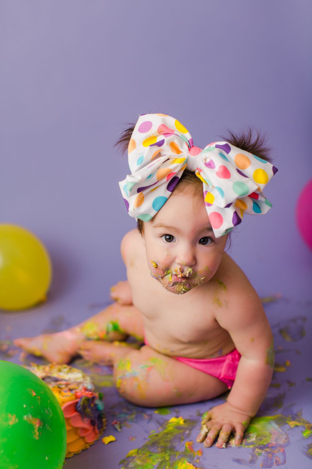 Baby with frosting-covered face and polka dot bow poses with smashed rainbow cake and balloons.