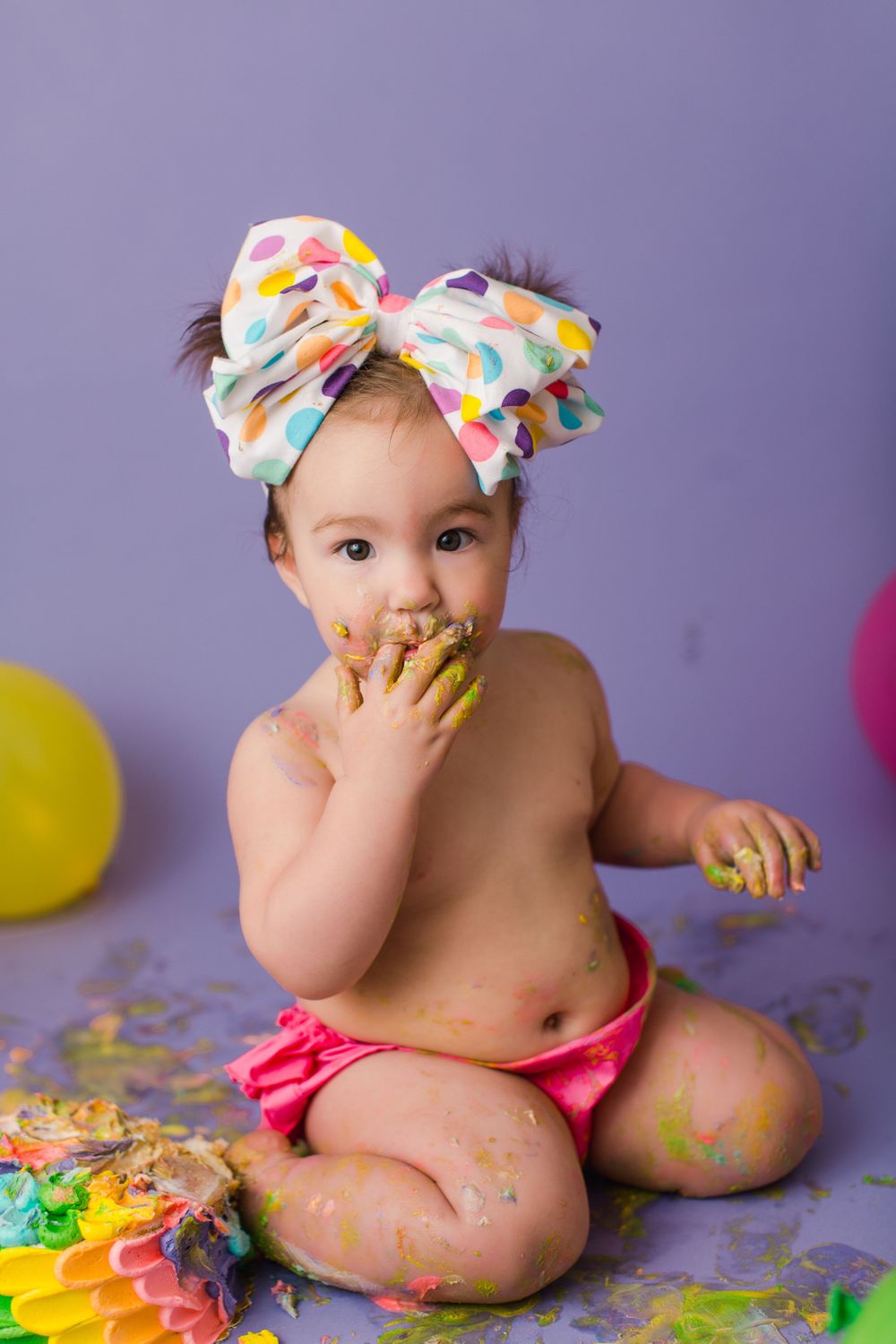 Baby in polka dot headband tastes colorful cake during festive cake smash photo session.