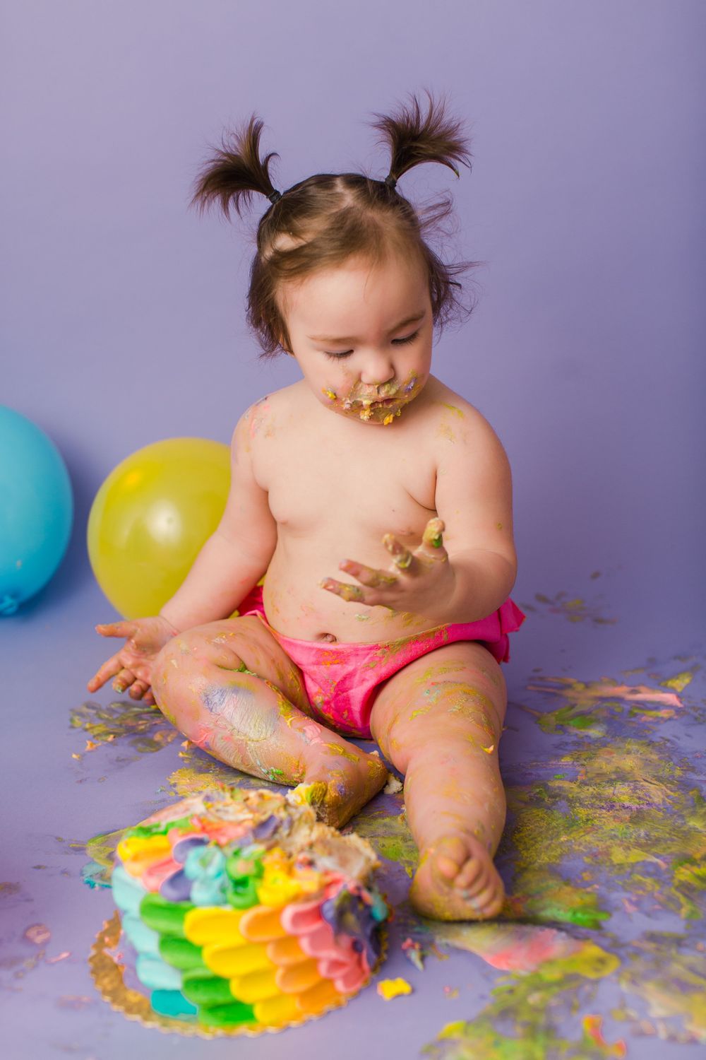 Baby with pigtails explores smashed rainbow cake during messy cake smash photography session.