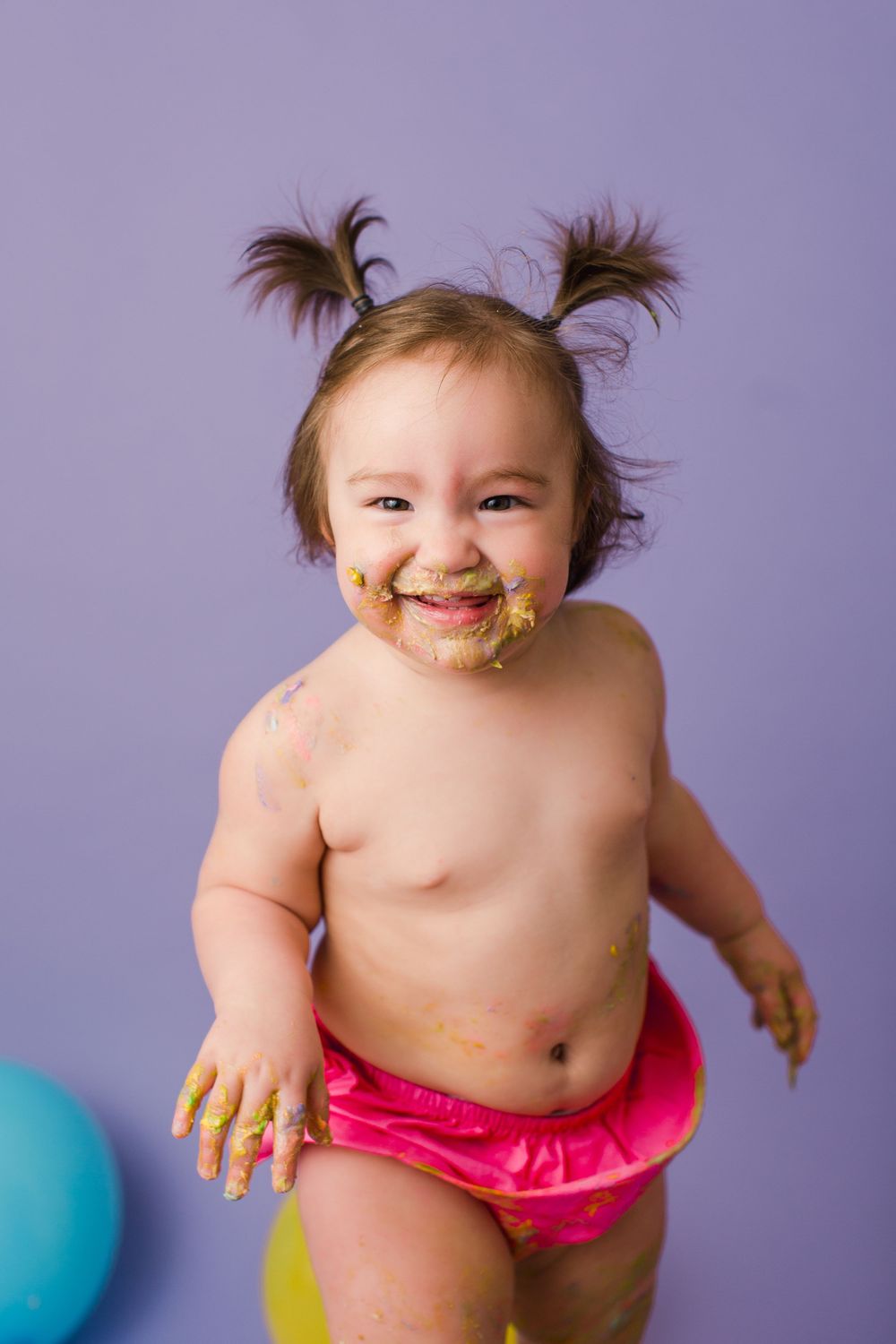Happy baby with messy face and pigtails plays during colorful cake smash photo session.