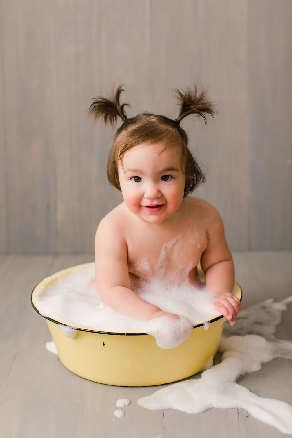 Happy baby with pigtails sits in vintage yellow basin filled with bubbles.