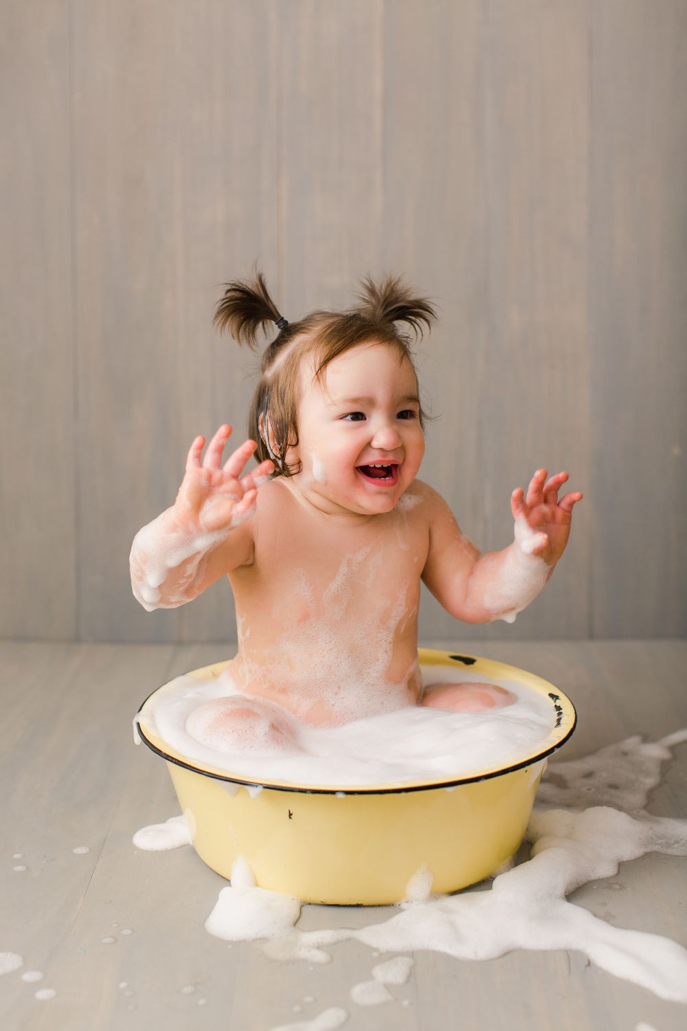 Happy baby splashing and playing in a yellow basin filled with bubbles and soap suds.
