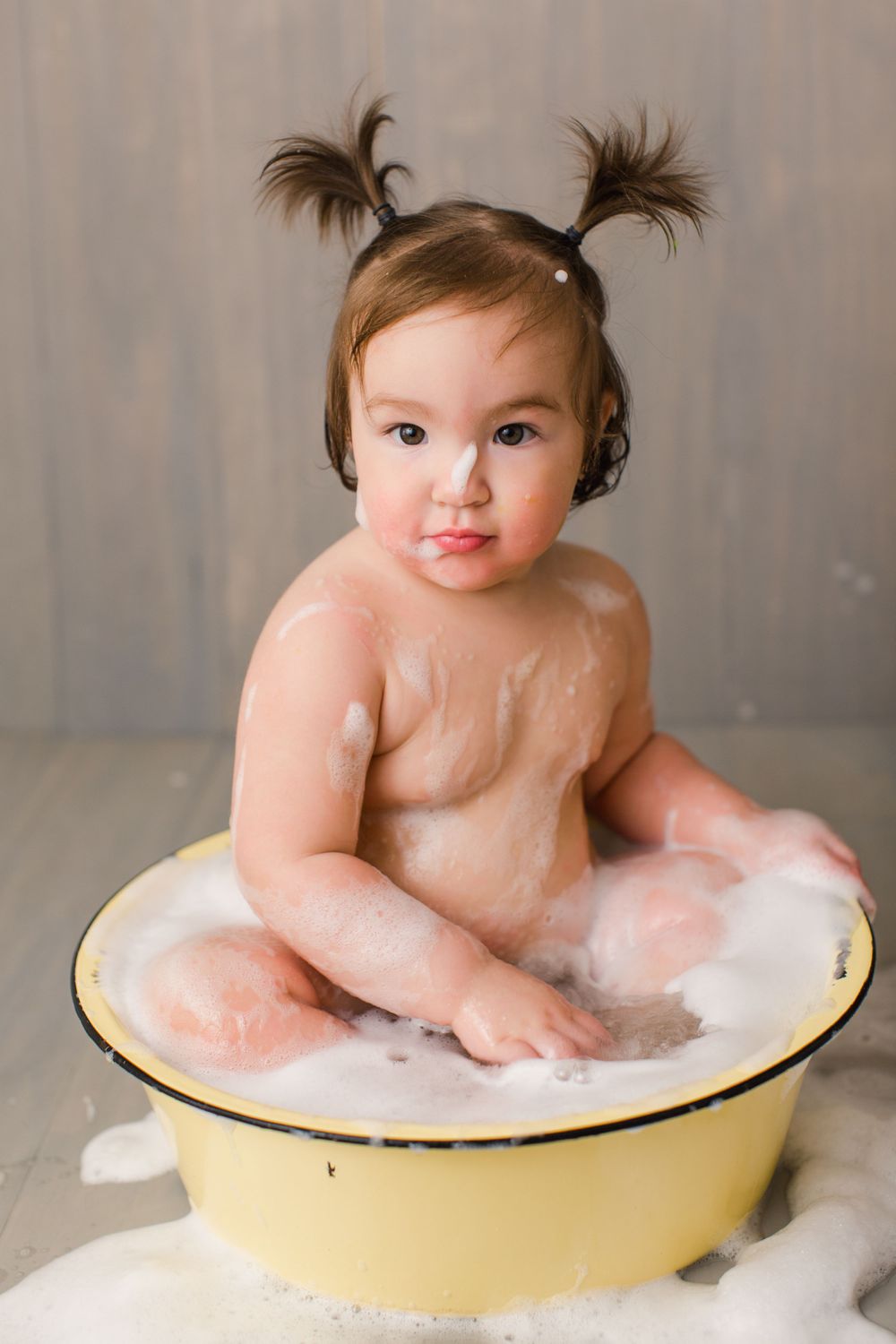 Baby with pigtails sitting in a yellow washbasin filled with soap bubbles.