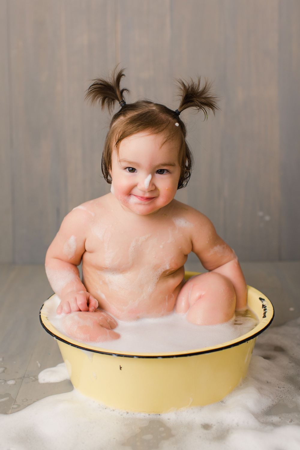 Joyful baby with spiky pigtails sitting in a yellow basin surrounded by bubbles.