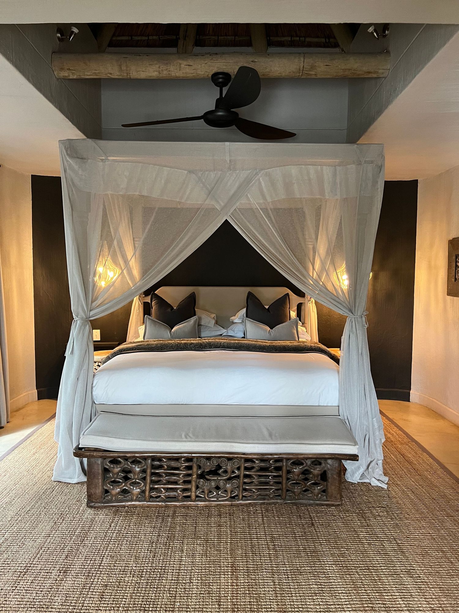 Elegant canopy bed with white draping and geometric bench in contemporary safari lodge bedroom.