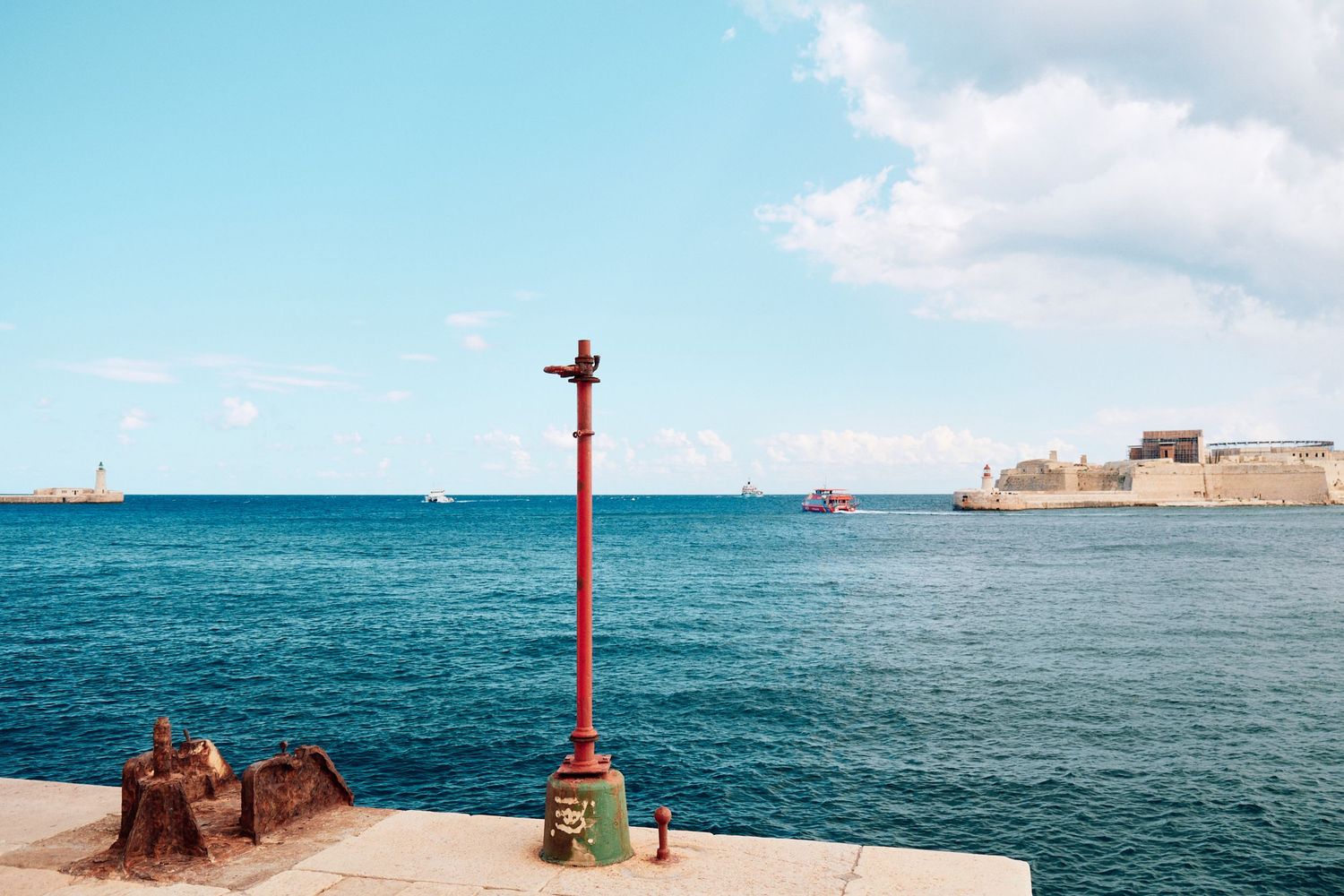 Red nautical pole stands against blue Mediterranean waters with fortress walls in background.