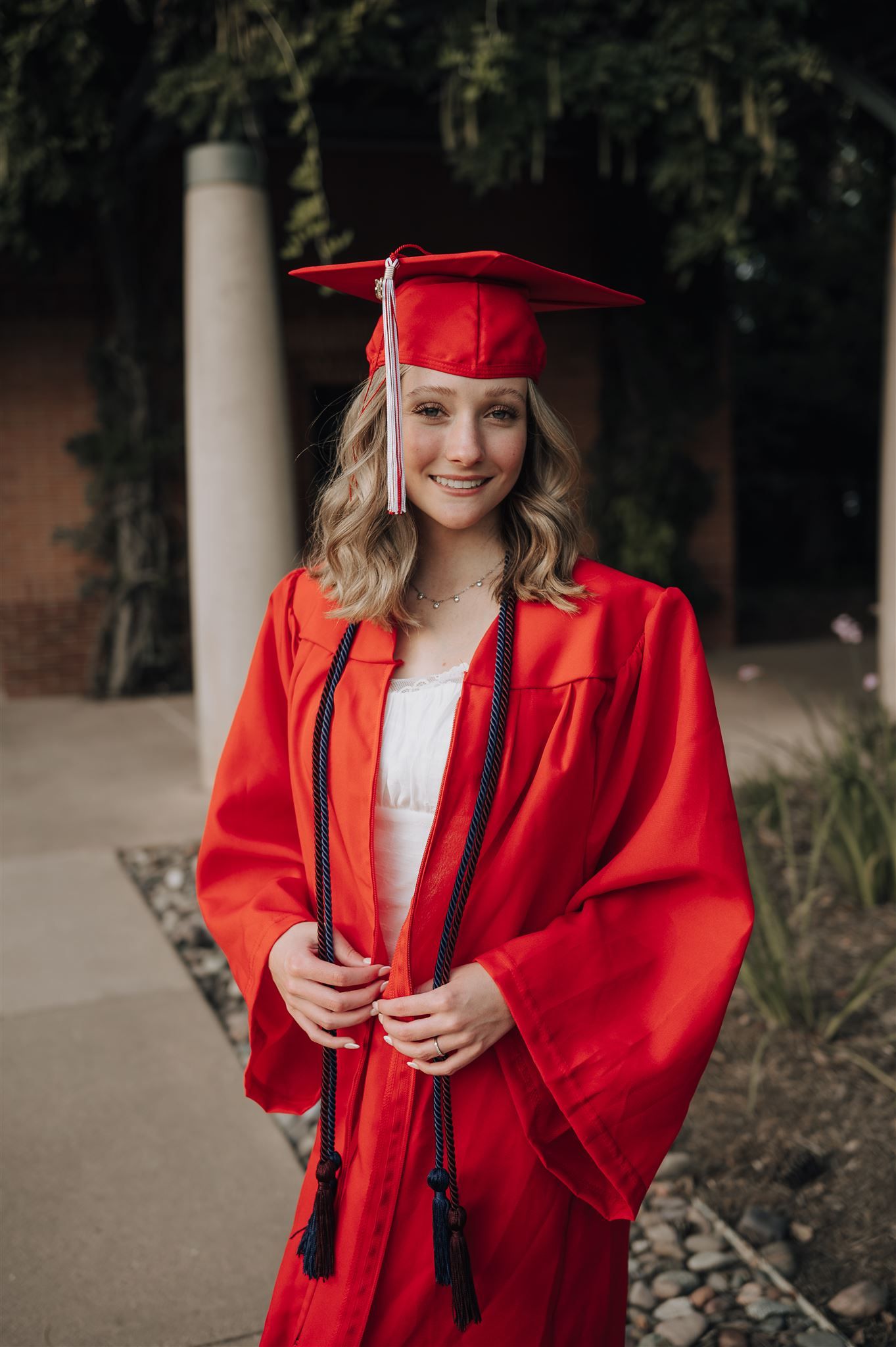 A graduate wearing a red cap and gown smiles while standing on a university campus pathway.