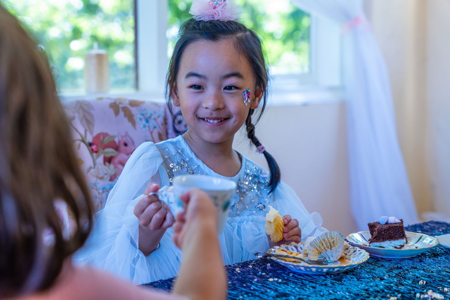 Young Asian girl in Elsa costume smiles while holding teacup at a princess birthday tea party.
