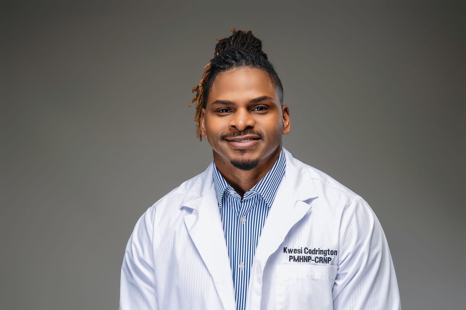 Healthcare professional in white coat and striped shirt with styled hair against gray background.