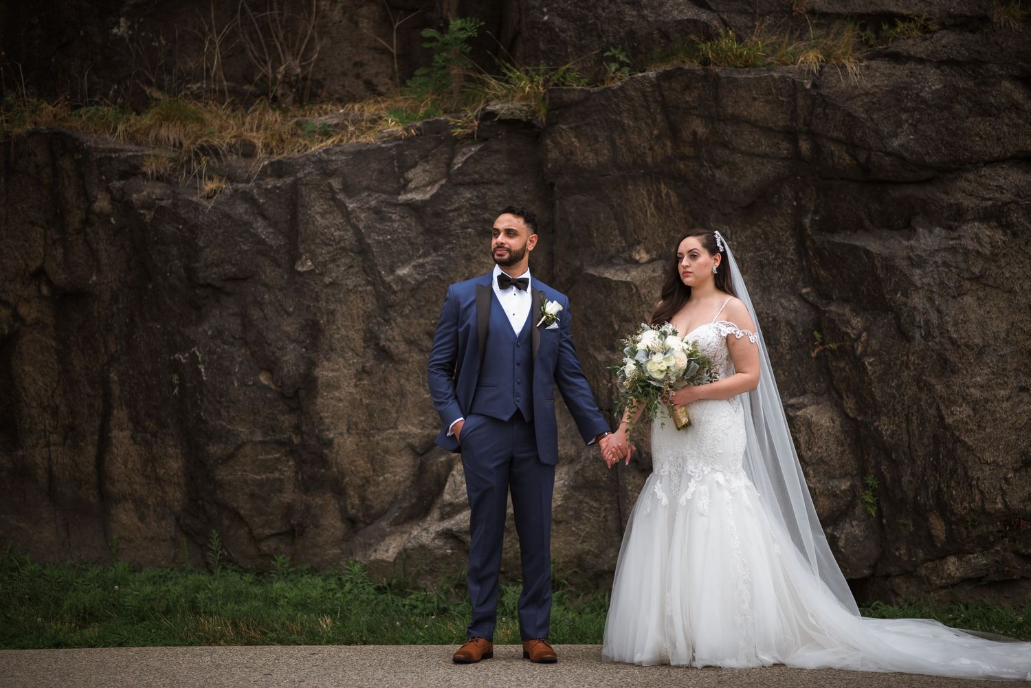 Bride and groom holding hands posing in front of a dramatic stone wall in a lush park setting.