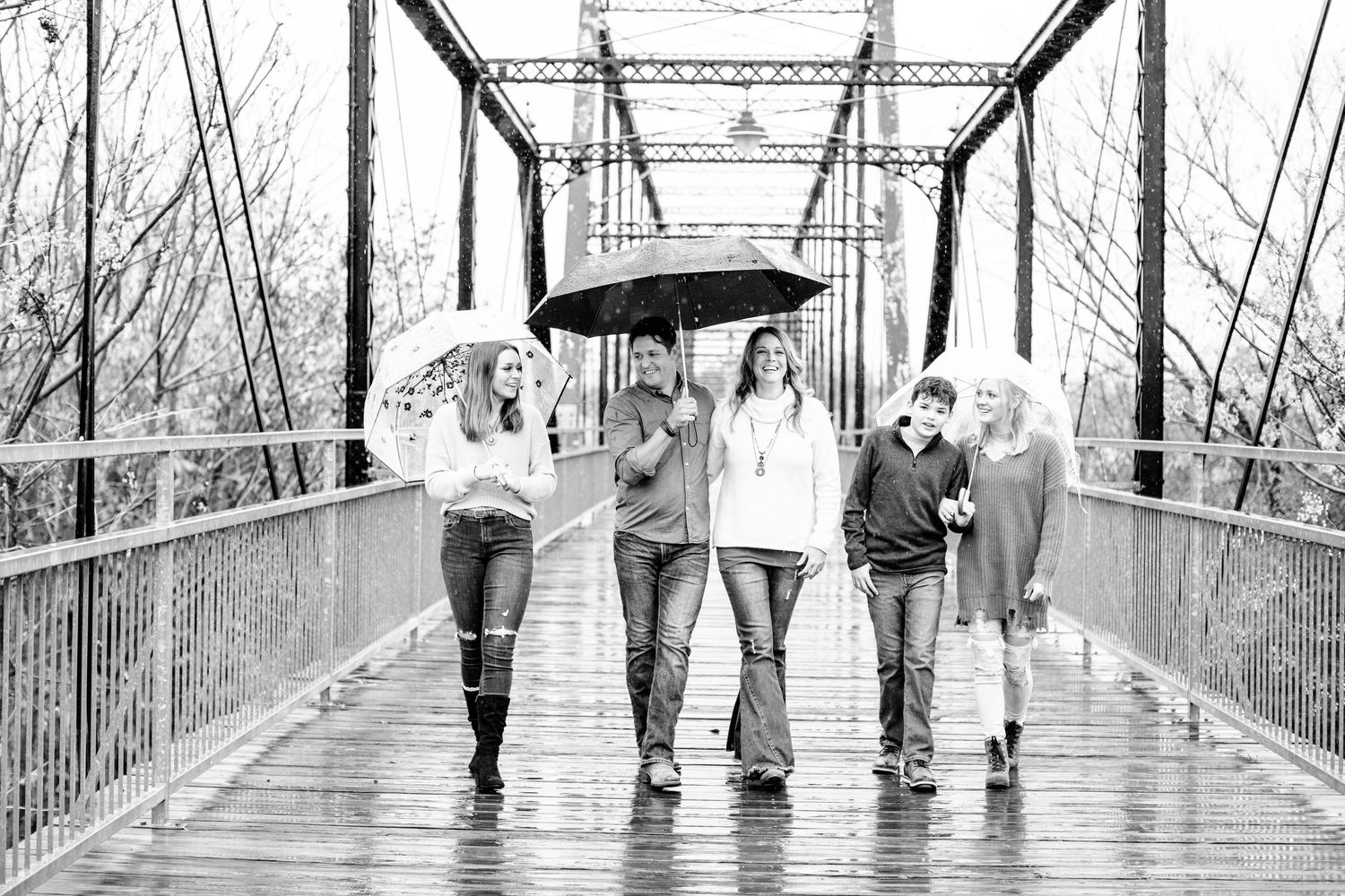 Group walking with umbrellas across rustic bridge in black and white photo.