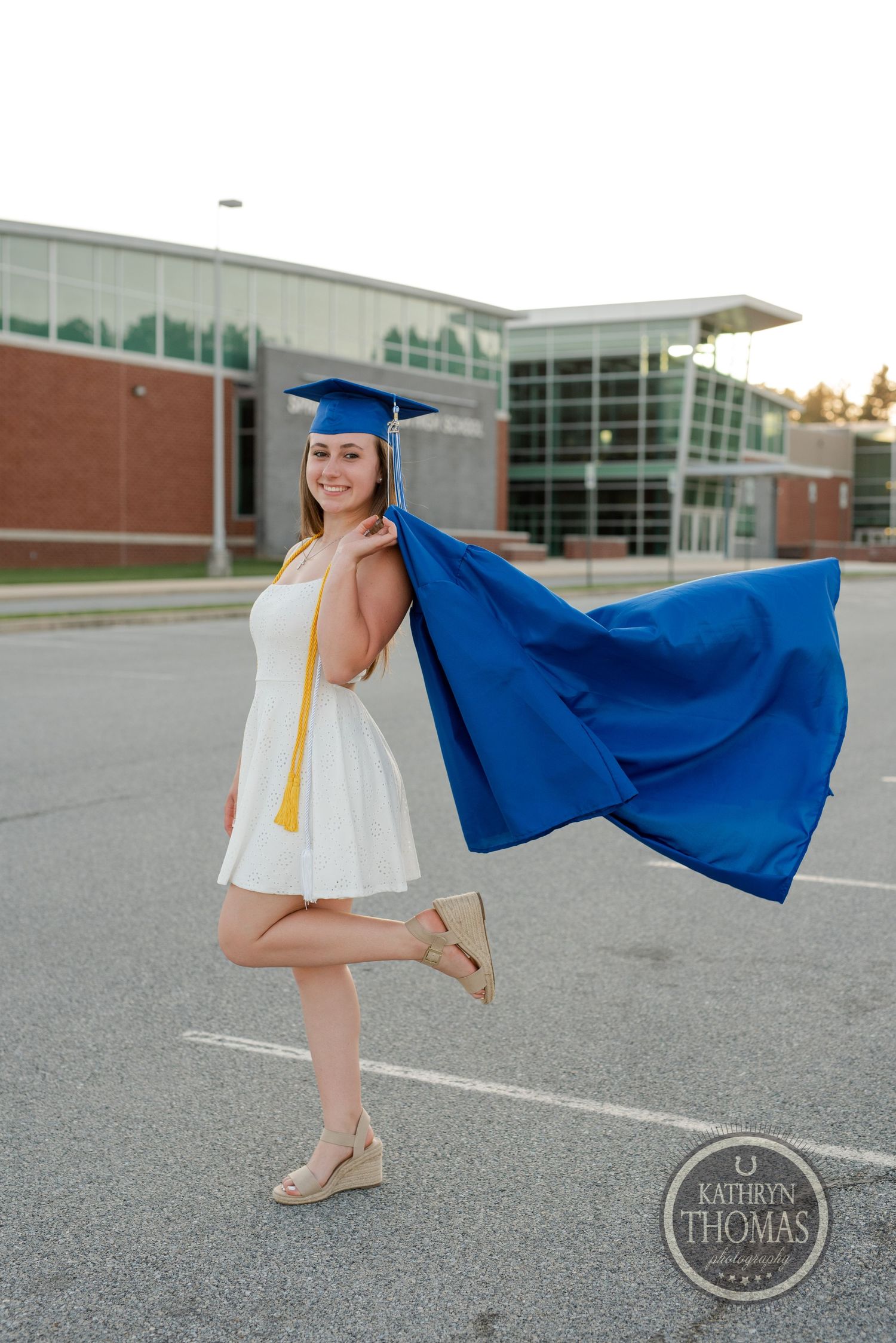 Graduate twirls in blue graduation gown and white dress in school parking lot.