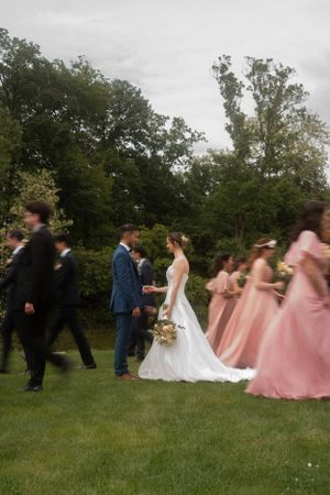 Wedding party walks across green lawn with bridesmaids in flowing pink dresses.