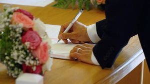 Close-up of hands signing marriage documents with pink flowers in the foreground.
