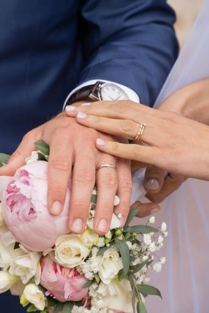 Close-up of newlywed couple's hands with wedding rings and pink floral bouquet.
