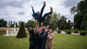 Wedding party jumps joyfully on a grassy lawn under cloudy skies.