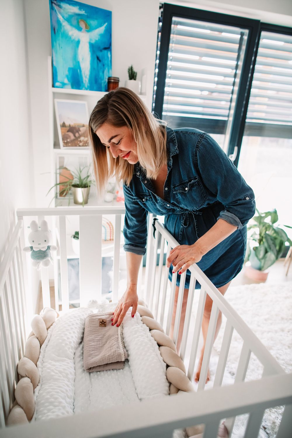 Expectant mother wearing denim dress arranges white bedding in a nursery crib near a blue wall art display.