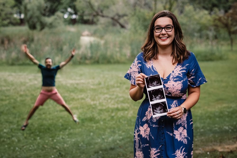 Woman in floral dress holding ultrasound photos while someone jumps joyfully in the background.