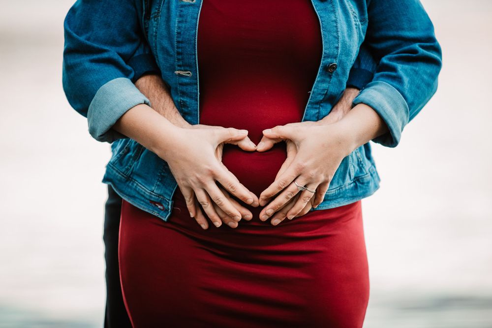 Close-up of hands forming heart shape over pregnant belly in denim jacket and red dress.