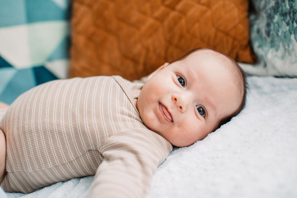 Baby in beige ribbed onesie lying on white bedding with colorful pillows in background.