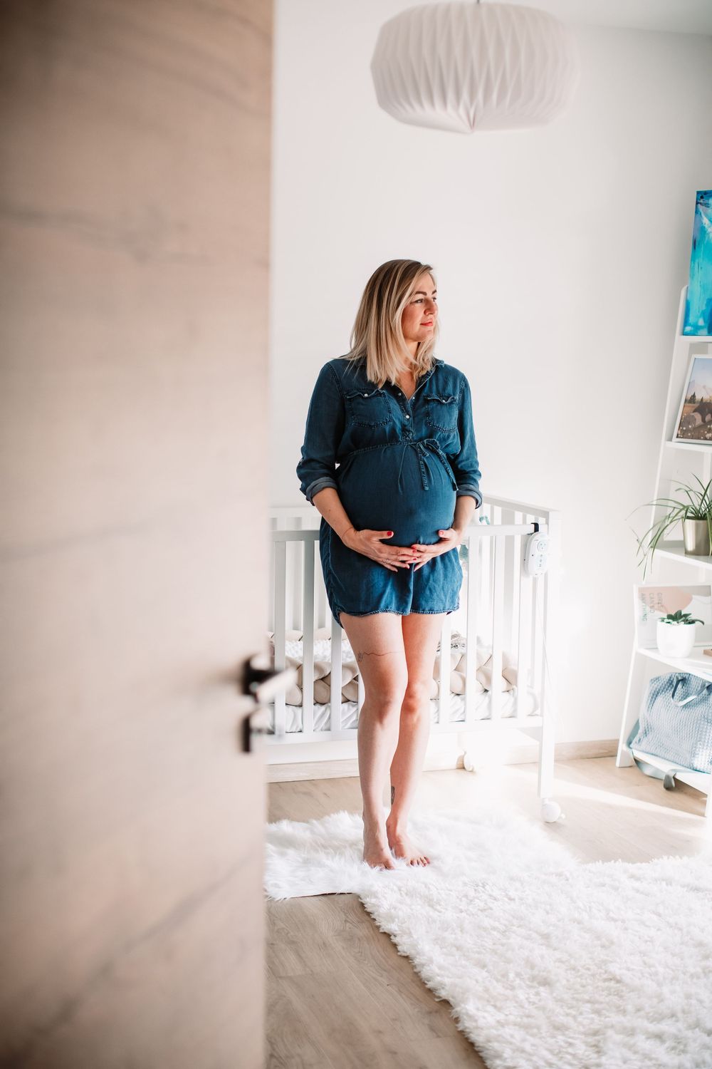 Expecting mother in blue denim dress stands by white crib in bright nursery with modern pendant light.