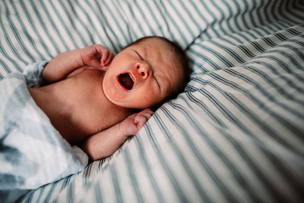 Sleepy newborn yawns while resting on blue and white striped bedding.