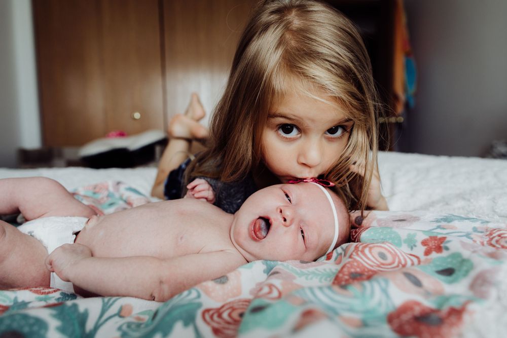 Toddler sibling lying next to newborn baby on floral bedding.
