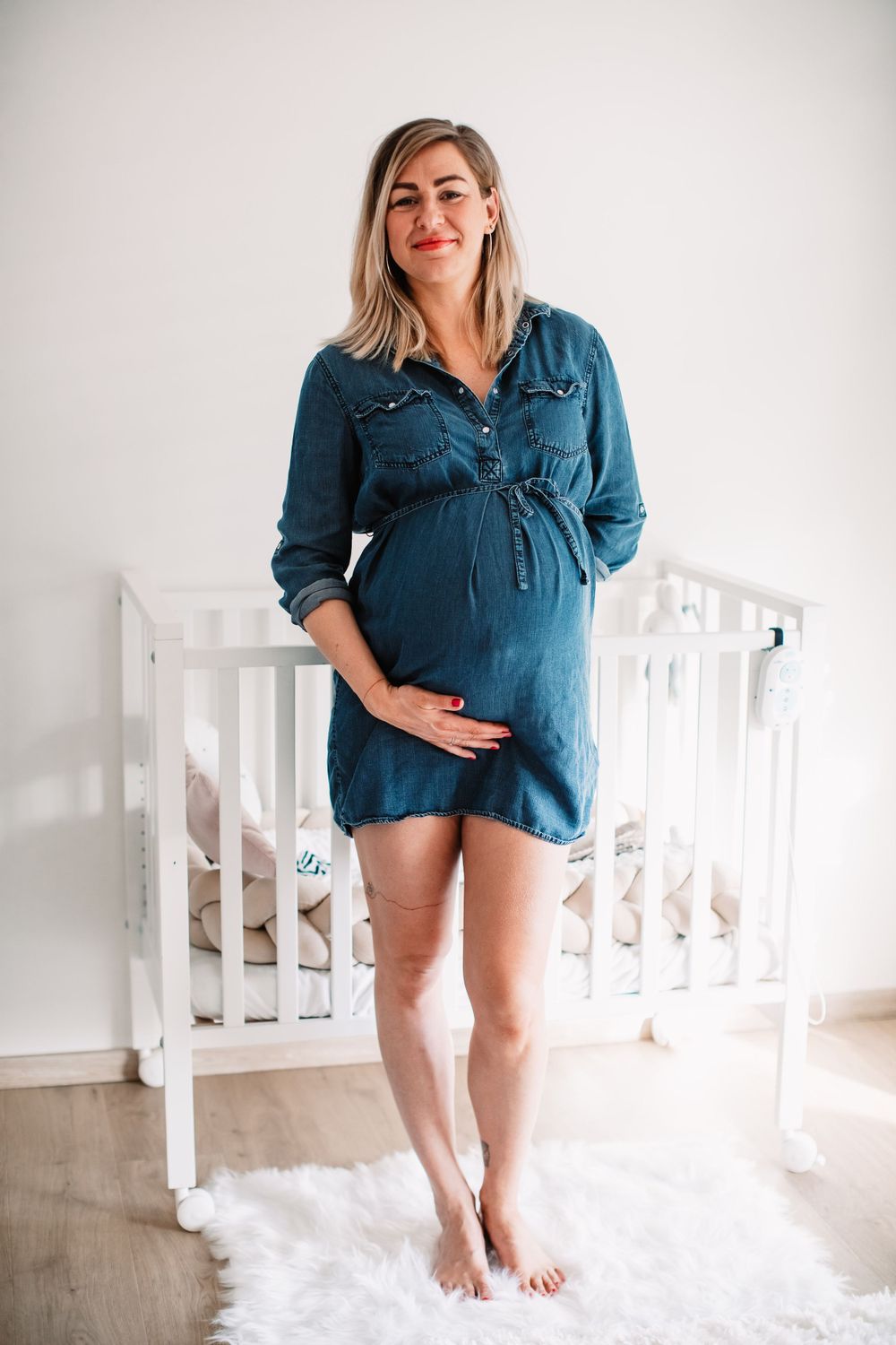 Pregnant woman in blue denim dress stands barefoot on white rug beside white crib in nursery.