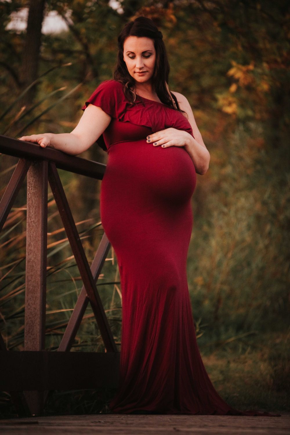 Maternity portrait of pregnant woman in flowing red dress standing on wooden bridge in autumn.
