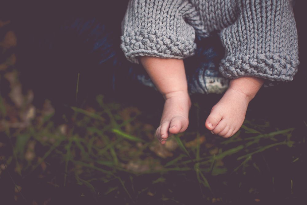 Tiny baby feet in gray knit pants against dark background with grass.