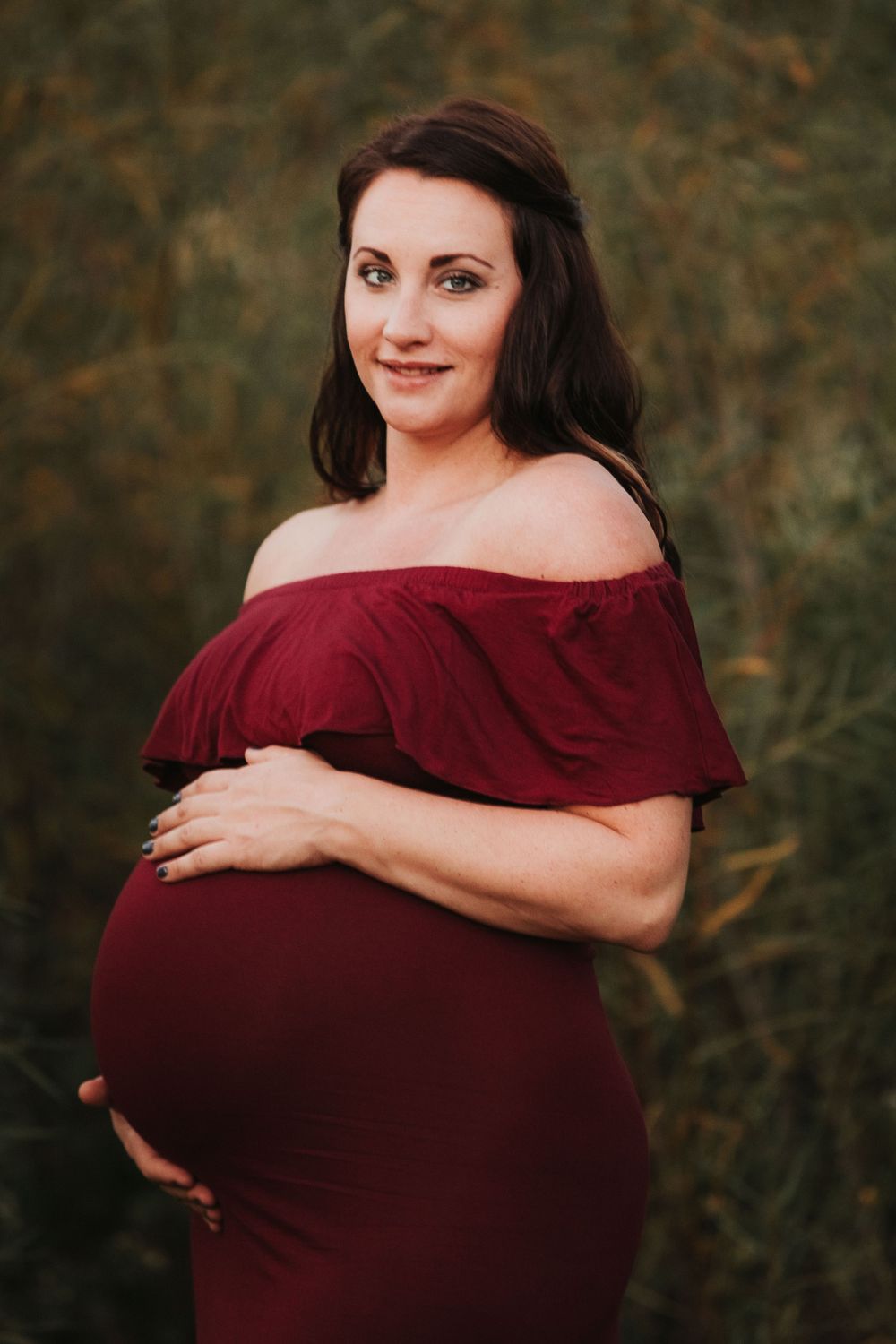 Maternity portrait featuring off-shoulder burgundy dress against natural outdoor backdrop.