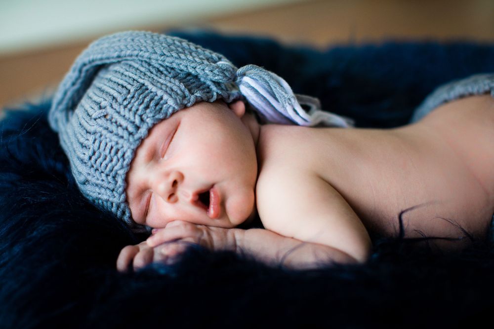 Sleeping baby wearing blue knitted hat on dark fabric.
