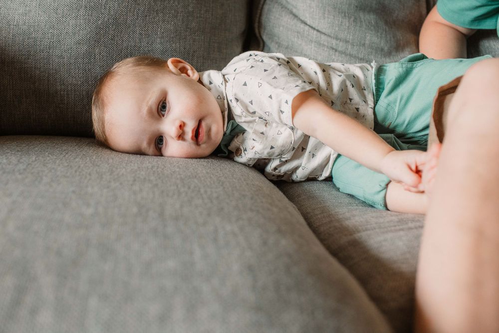 Young child rests peacefully on a gray couch wearing a patterned shirt and mint green pants.