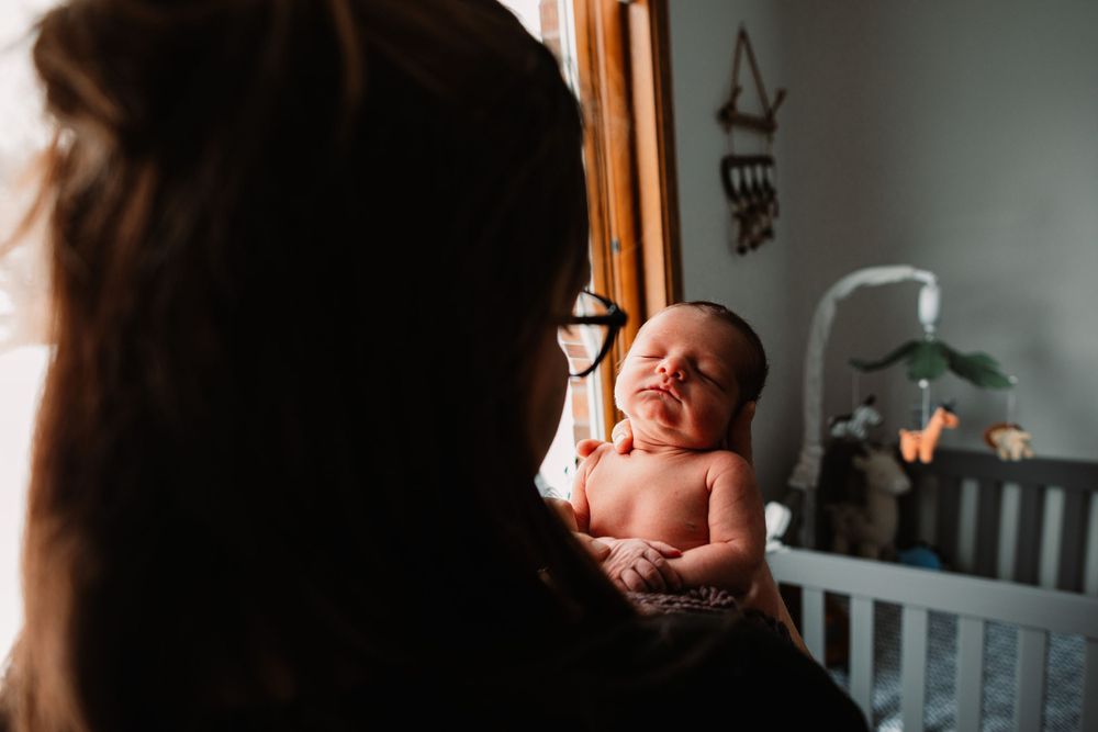 Newborn baby stretches while being held in a nursery with a crib visible in background.