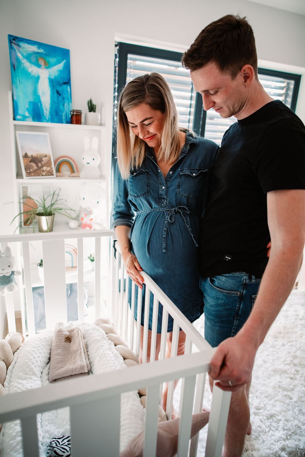 Couple looking into white nursery crib with decorative wall shelves and blue artwork in background.