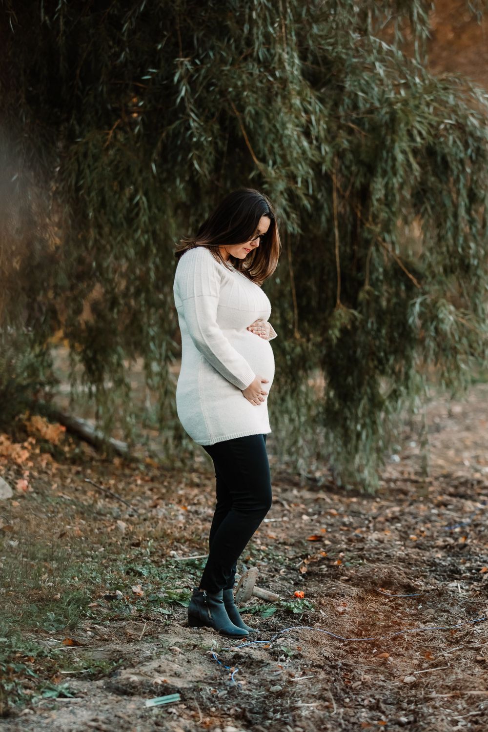 Side view of pregnant woman in white sweater standing against willow tree background.
