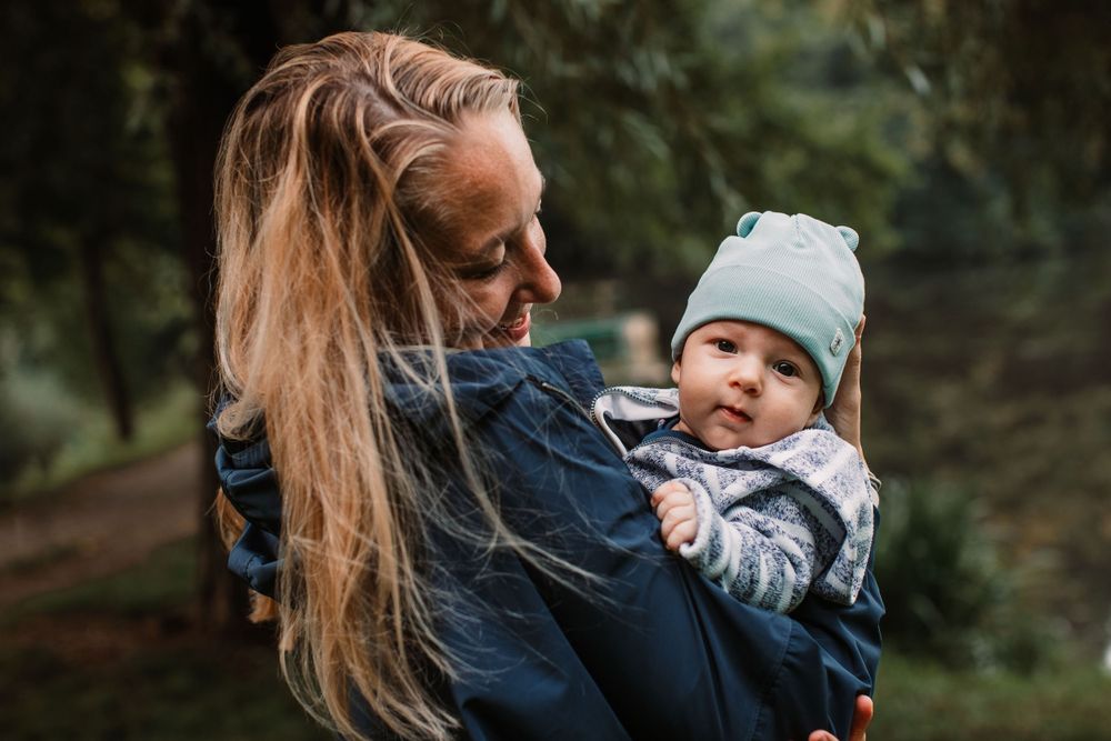 A parent holds a bundled baby wearing a mint beanie while walking outdoors in nature.