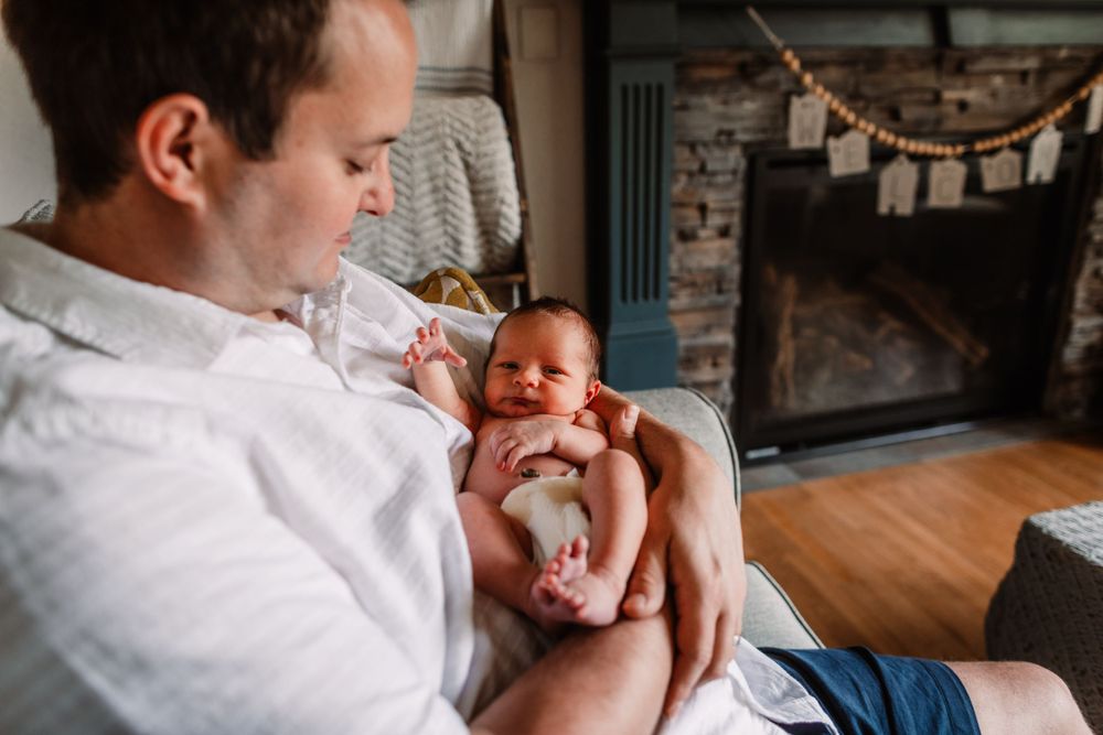 Parent holding newborn baby in white shirt against rustic indoor setting with string lights.