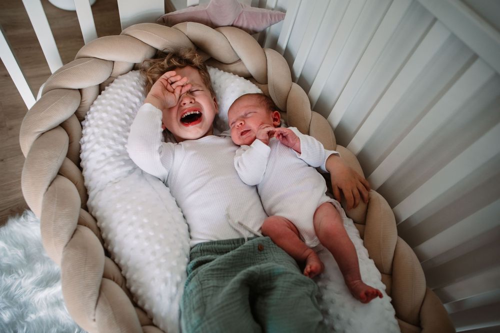 Two children laying on braided cushion in crib sharing laughter.