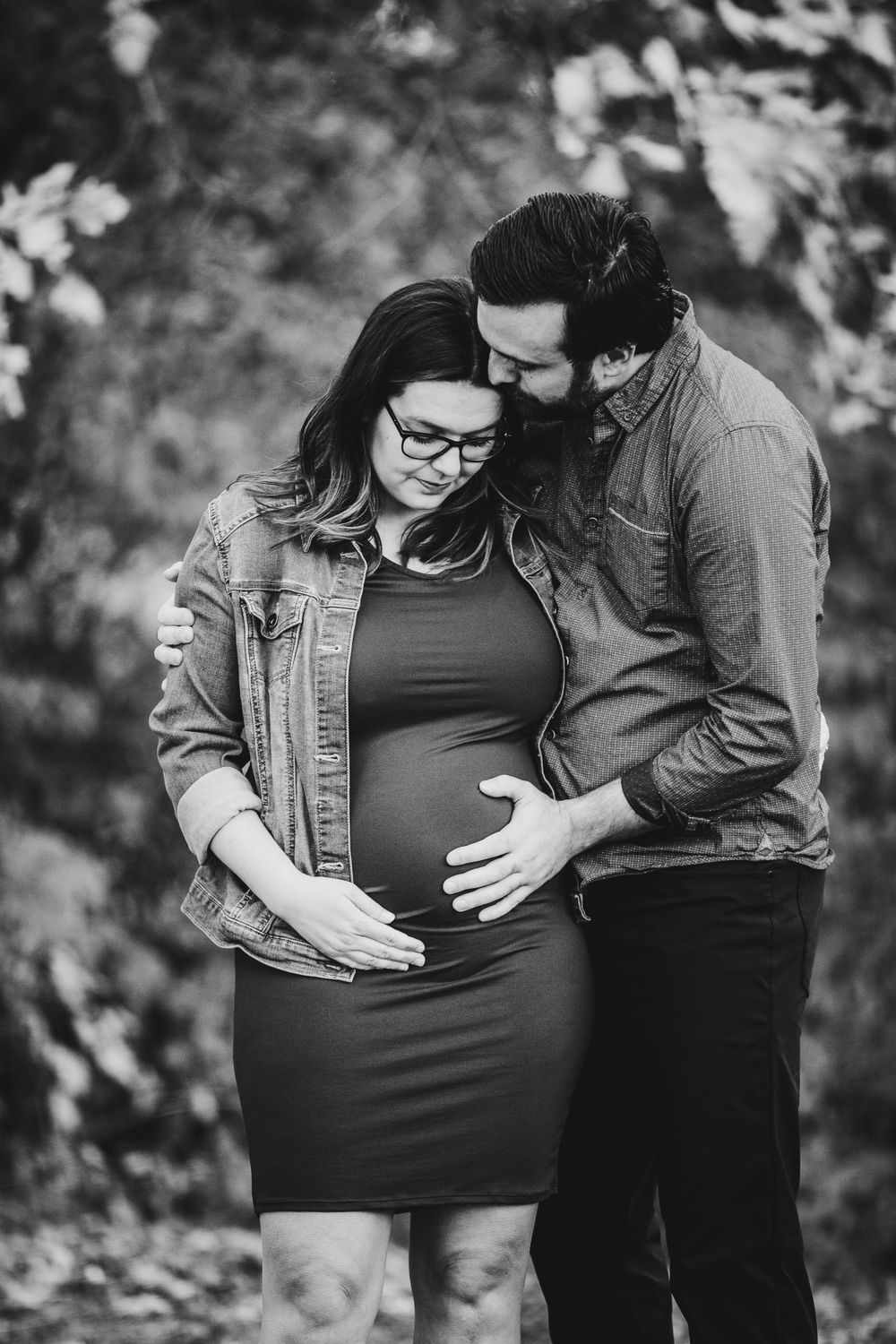 Black and white maternity photo of expectant couple embracing outdoors with natural foliage in background.