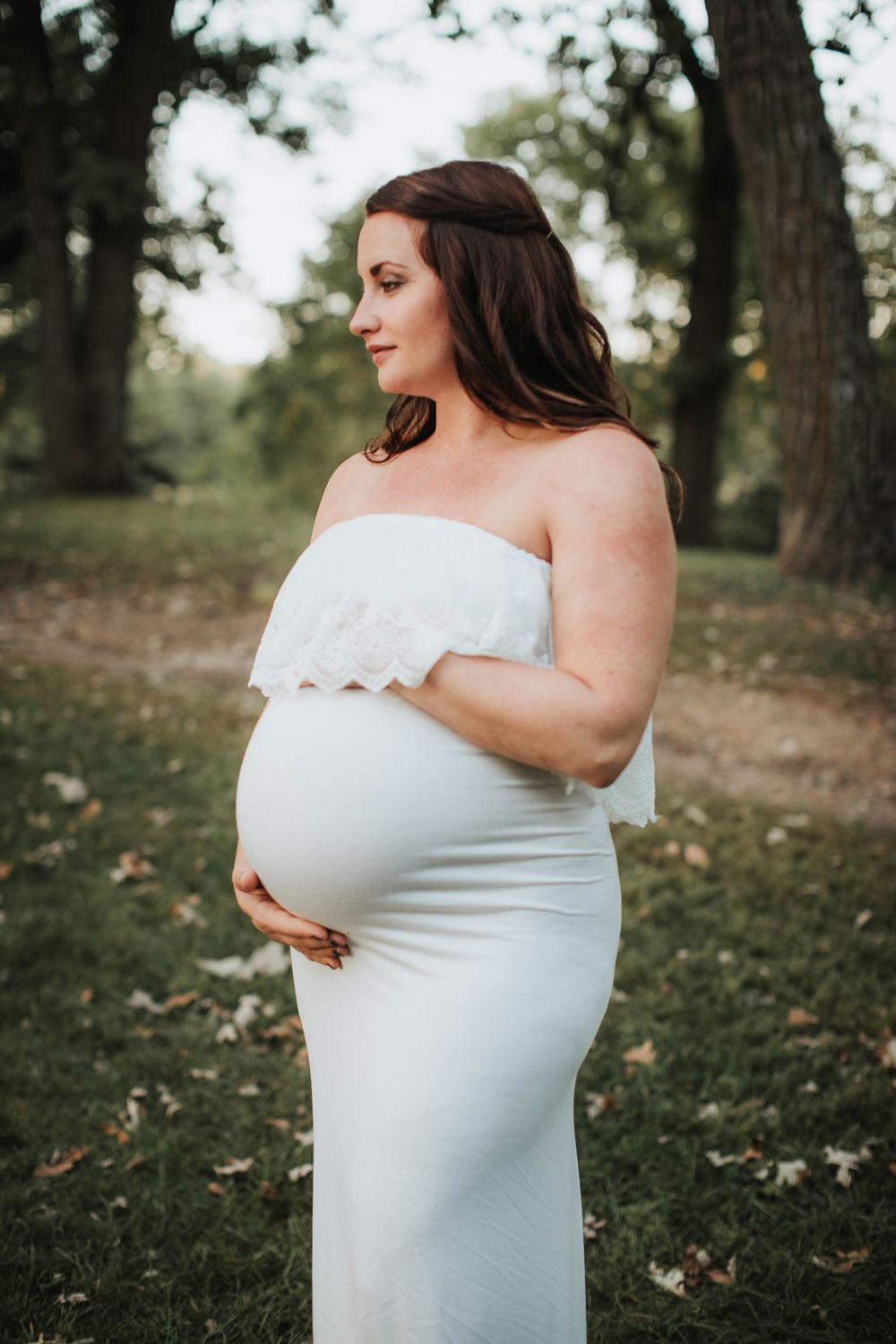 Elegant maternity portrait in white off-shoulder dress against green park background.