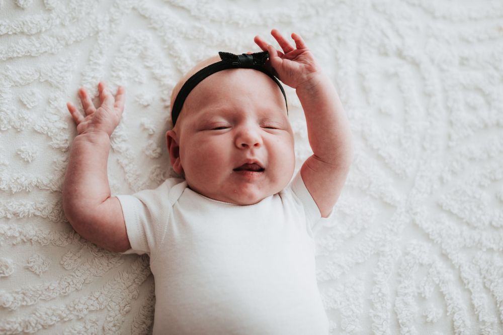 Sleeping newborn wearing white onesie and headband on white blanket.