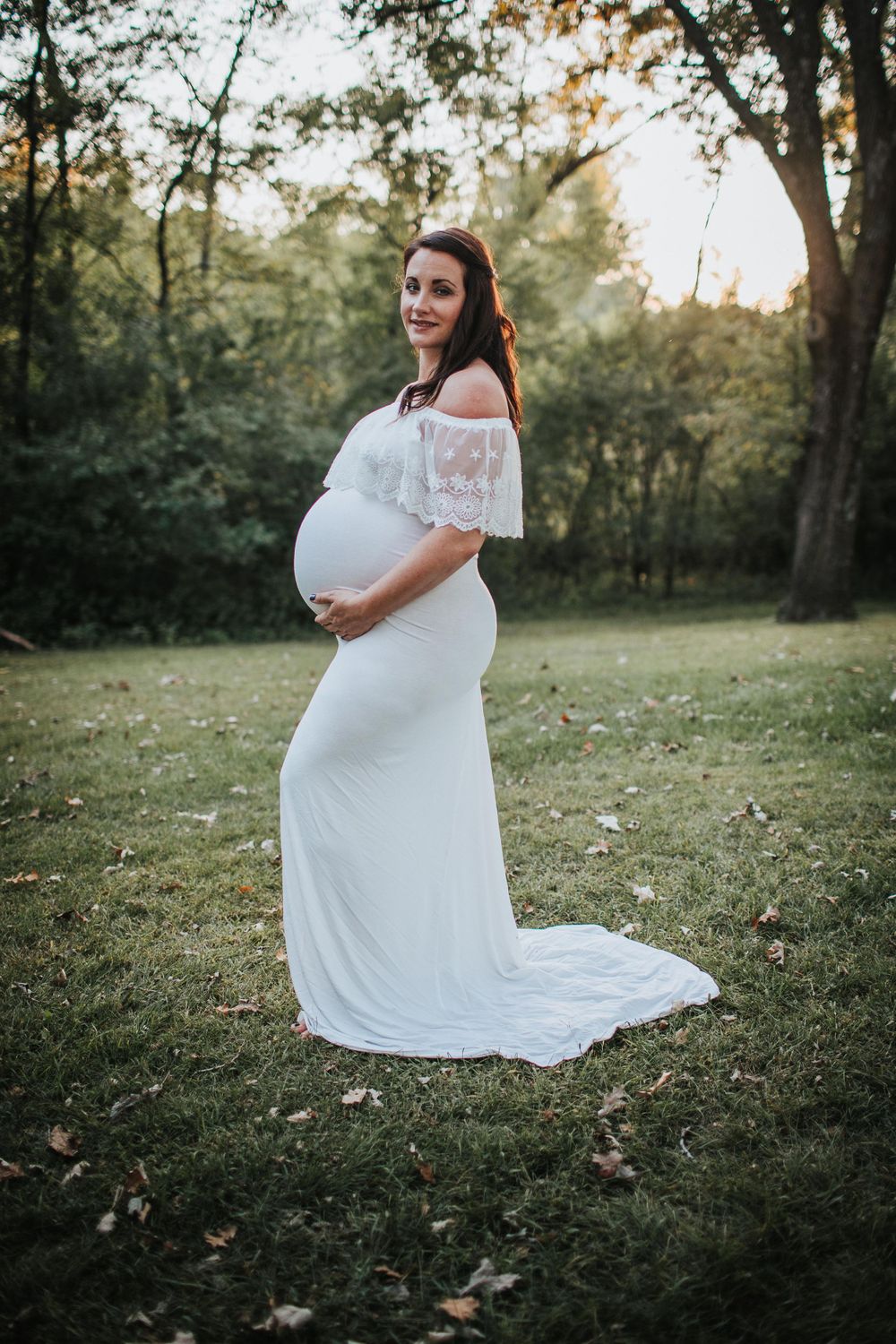 Outdoor maternity photo featuring white lace gown and natural wooded setting at sunset.