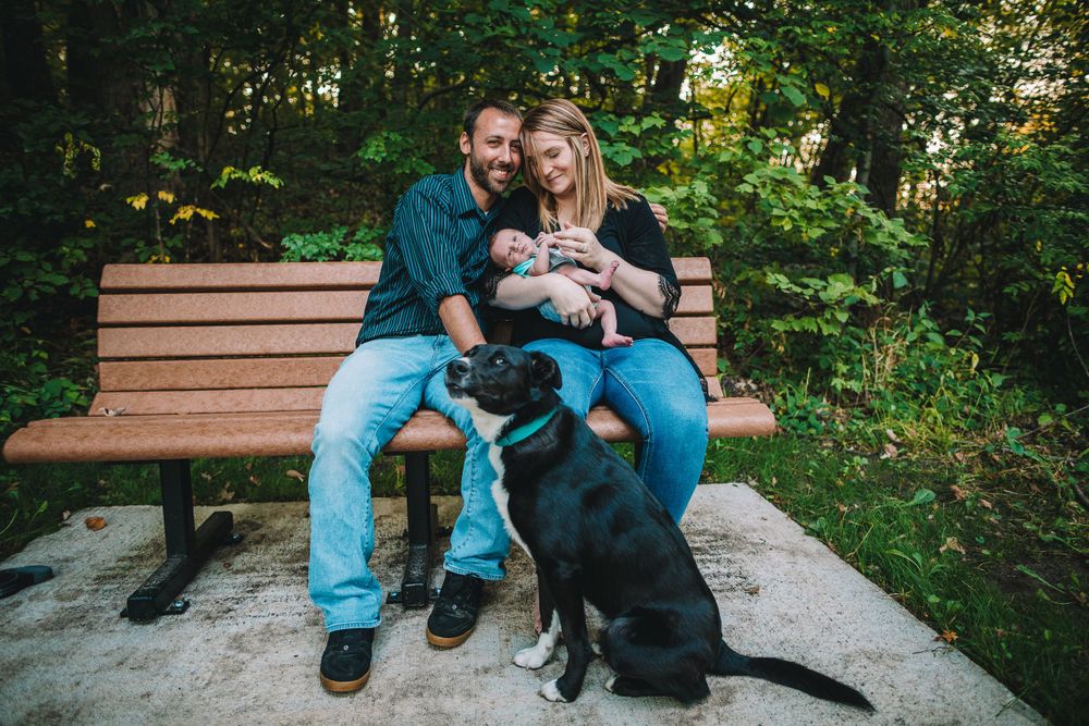 Family sitting on park bench with baby and black dog in natural setting.