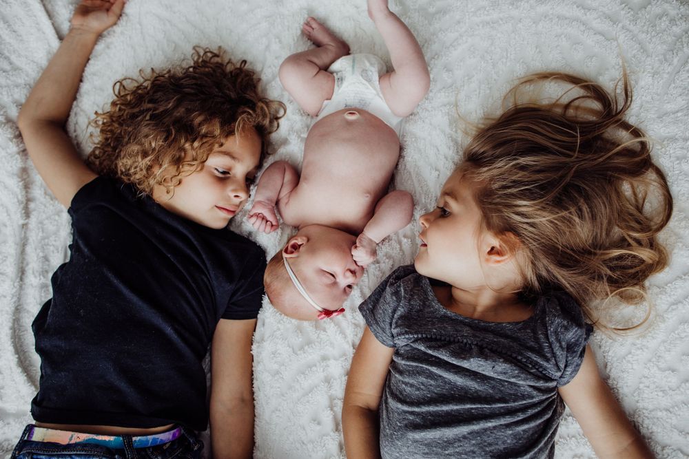 Overhead view of siblings lying together with newborn on white bedding.