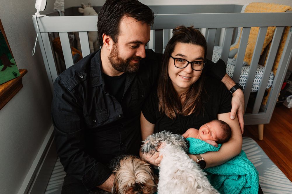 Couple sitting in nursery with baby and small white dog near gray crib.