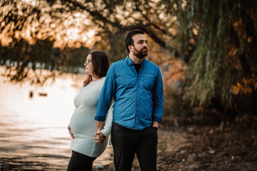 Couple standing back-to-back during maternity photoshoot with sunset lighting through trees.