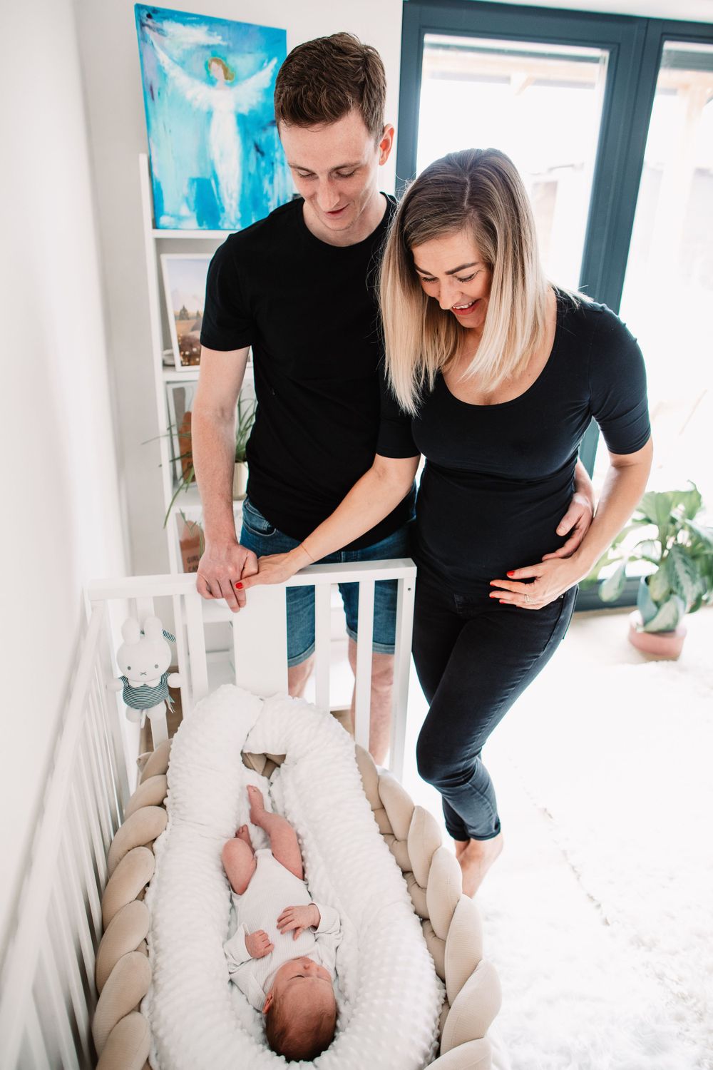 Parents looking down lovingly at their newborn baby sleeping in a white cushioned crib.